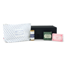Produits du coffret bien-être posés devant la boîte noire : pochette Puy du Fou 1900 bleue, lavant floral solide, savon rose et déodorant solide coton.