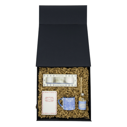 Coffret Maison & Senteurs ouvert vu du dessus : bougies parfumées, carnet A6, mini mug bleu et diffuseur disposés dans les frisures.