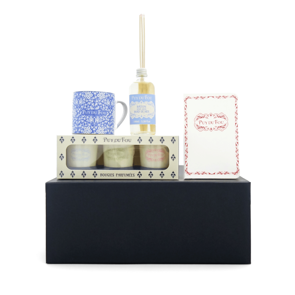 Coffret Maison & Senteurs Puy du Fou posé sur la boîte : trois bougies parfumées, mini mug bleu, diffuseur senteur bord de mer et carnet A6 1900.